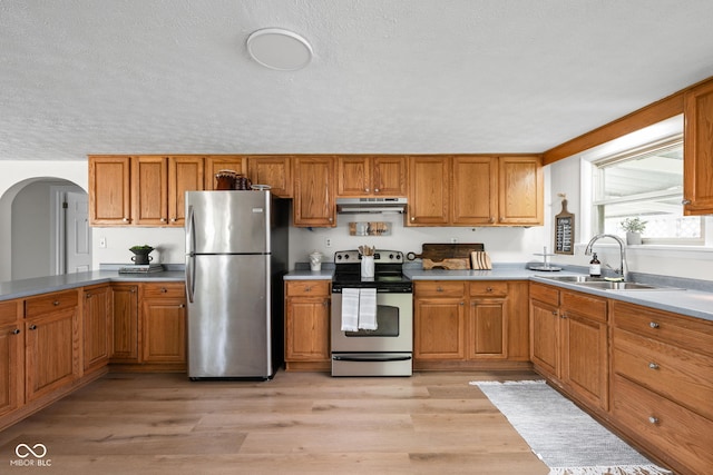 kitchen with appliances with stainless steel finishes, under cabinet range hood, a sink, arched walkways, and brown cabinets