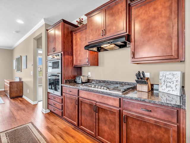 kitchen featuring dark stone countertops, light wood-style flooring, stainless steel appliances, under cabinet range hood, and crown molding