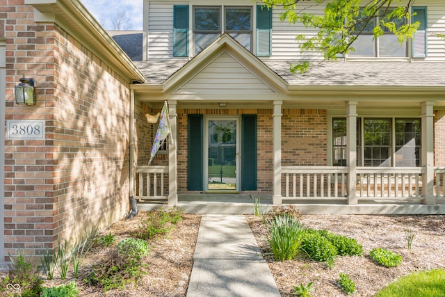 view of exterior entry featuring covered porch, a shingled roof, and brick siding
