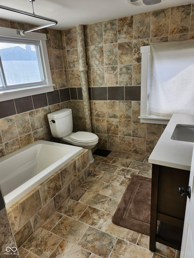 bathroom featuring tiled bath, vanity, toilet, stone tile floors, and tile walls