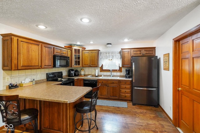 kitchen with brown cabinetry, a sink, black appliances, a peninsula, and dark wood-style floors