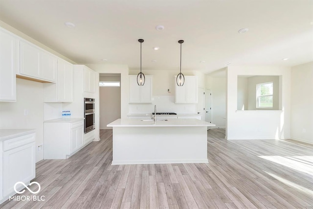 kitchen with stainless steel double oven, an island with sink, light wood-style flooring, white cabinetry, and light countertops
