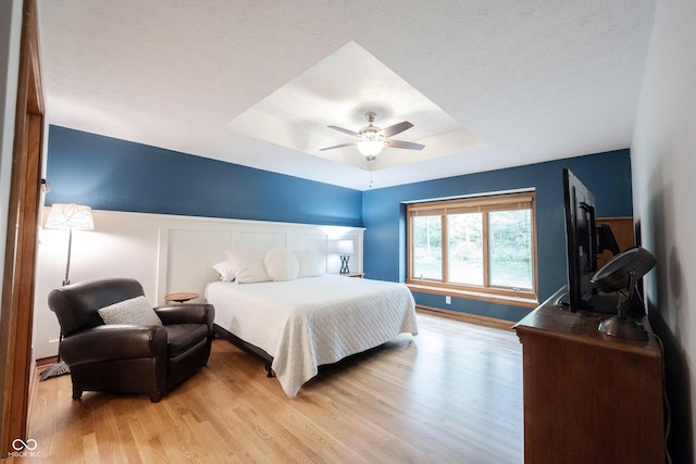 bedroom with a wainscoted wall, a tray ceiling, ceiling fan, and light wood-style flooring