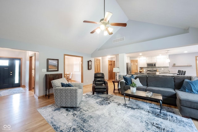 living room featuring light wood-style flooring, baseboards, ceiling fan, visible vents, and high vaulted ceiling