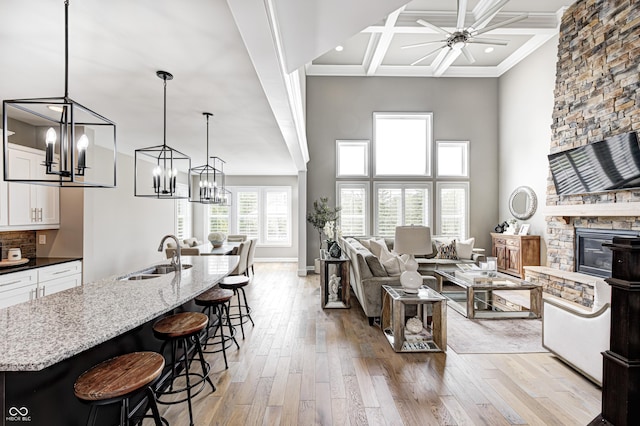 living area with light wood finished floors, a stone fireplace, ceiling fan with notable chandelier, recessed lighting, and coffered ceiling