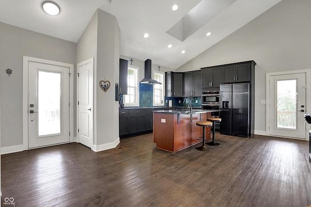 kitchen featuring wall chimney range hood, a kitchen island with sink, dark wood-style flooring, refrigerator with ice dispenser, and a breakfast bar