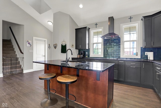 kitchen with dark wood-type flooring, dark stone counters, wall chimney exhaust hood, a sink, and backsplash