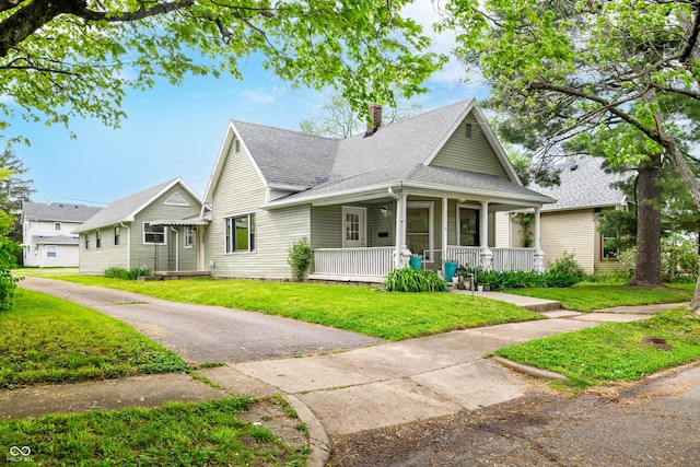 bungalow-style home with roof with shingles, covered porch, a chimney, and a front lawn