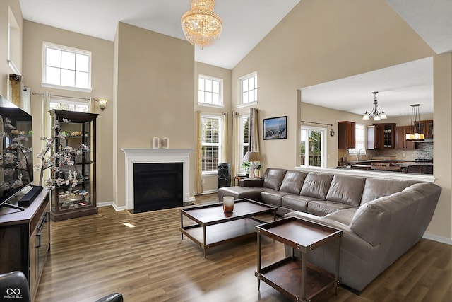 living area featuring a chandelier, high vaulted ceiling, wood finished floors, a fireplace with flush hearth, and baseboards