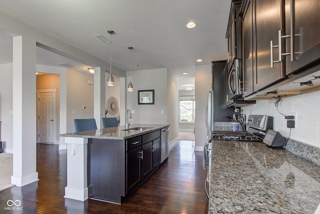 kitchen featuring appliances with stainless steel finishes, a center island with sink, a sink, dark stone countertops, and dark wood-type flooring