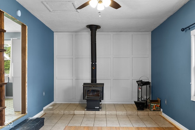 unfurnished living room with a decorative wall, baseboards, ceiling fan, a wood stove, and light tile patterned flooring