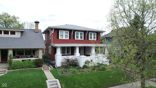 view of front of home with a shingled roof, a front yard, covered porch, and a chimney
