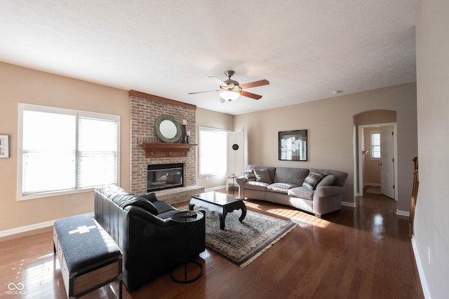 living area featuring ceiling fan, arched walkways, a brick fireplace, wood finished floors, and baseboards