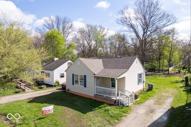 bungalow featuring covered porch, a front yard, cooling unit, roof with shingles, and driveway