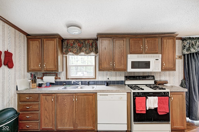 kitchen with a sink, white appliances, a textured ceiling, brown cabinetry, and wallpapered walls