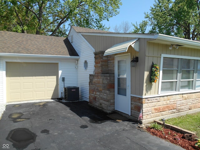 view of side of home featuring roof with shingles, central AC unit, driveway, and stone siding