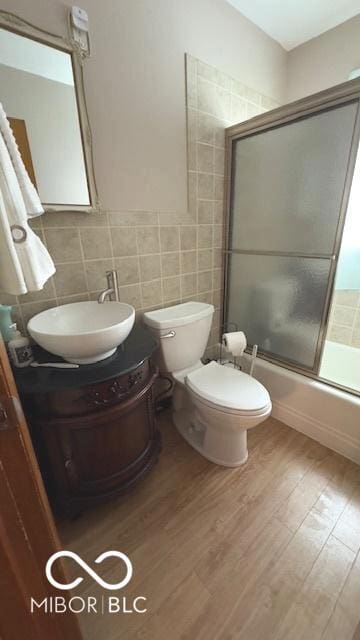bathroom with vanity, toilet, tile walls, wood finished floors, and bath / shower combo with glass door