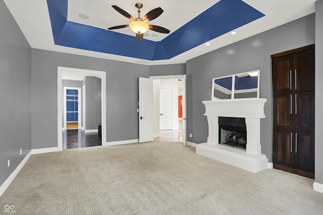 unfurnished living room featuring a ceiling fan, carpet floors, baseboards, a fireplace with raised hearth, and a tray ceiling
