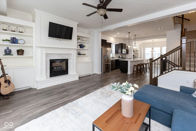 living room with built in features, a fireplace, ceiling fan with notable chandelier, stairs, and dark wood-type flooring