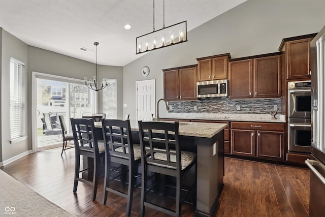 kitchen featuring backsplash, dark wood-style flooring, an inviting chandelier, stainless steel appliances, and an island with sink