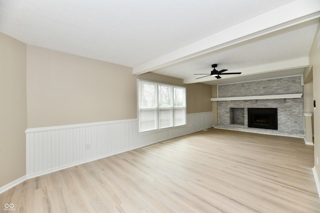 unfurnished living room featuring wainscoting, beam ceiling, light wood-style floors, a large fireplace, and a ceiling fan