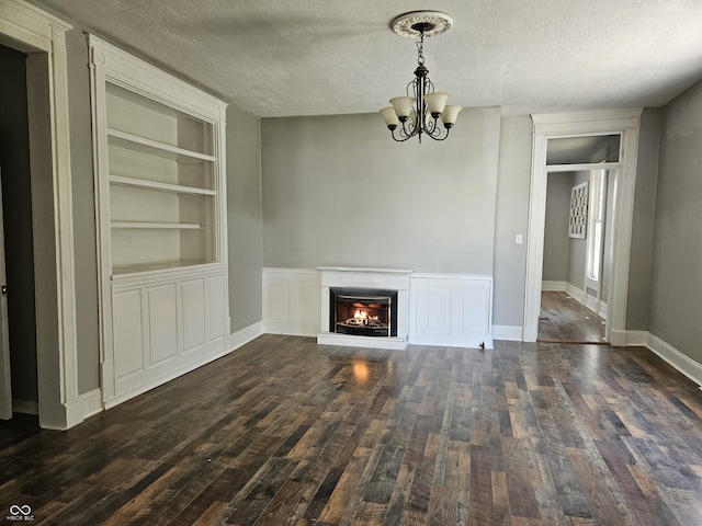 unfurnished living room featuring an inviting chandelier, a textured ceiling, built in features, dark wood finished floors, and a glass covered fireplace