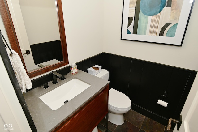 bathroom featuring toilet, stone finish floor, vanity, and a wainscoted wall