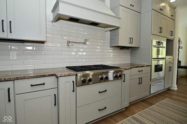 kitchen featuring stainless steel appliances, dark wood-style flooring, a warming drawer, decorative backsplash, and premium range hood