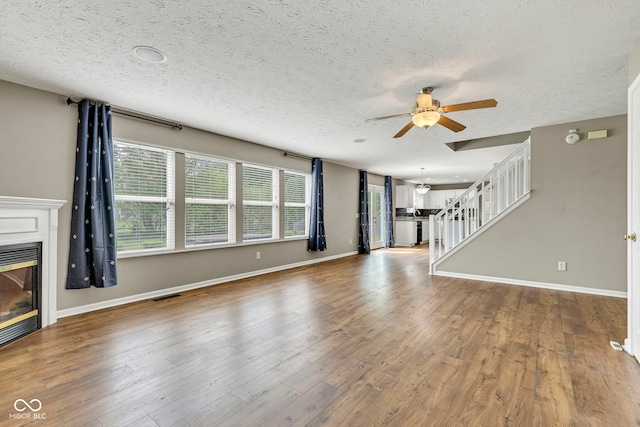 unfurnished living room with a ceiling fan, a textured ceiling, a glass covered fireplace, visible vents, and stairs