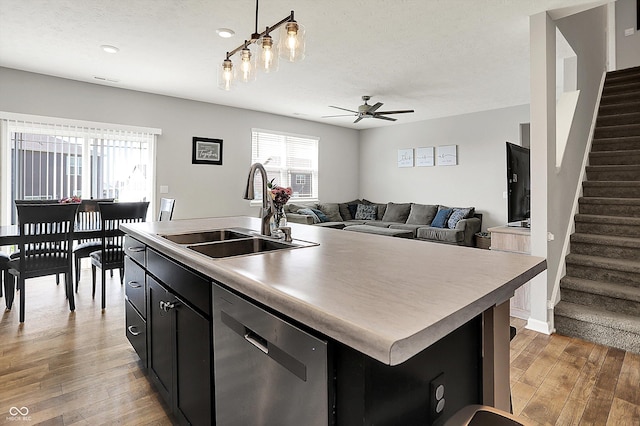 kitchen with a kitchen island with sink, stainless steel dishwasher, a sink, dark cabinetry, and light wood-style flooring