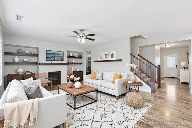 living area featuring a ceiling fan, wood finished floors, a glass covered fireplace, stairway, and built in features