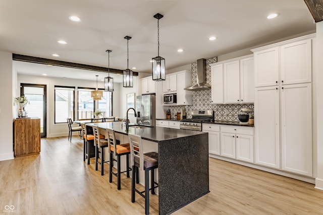1534 Fletcher Ave, Indianapolis, IN 46203 - kitchen featuring wall chimney range hood, appliances with stainless steel finishes, a center island with sink, light wood-style floors, and dark countertops