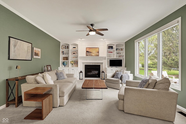carpeted living room with a ceiling fan, built in features, a fireplace, baseboards, and ornamental molding