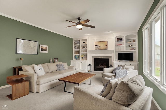 carpeted living room featuring built in features, a ceiling fan, baseboards, a tile fireplace, and ornamental molding