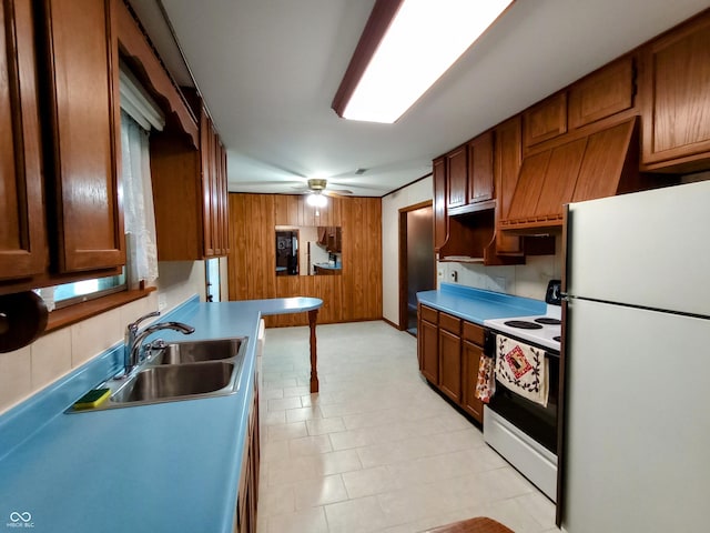 kitchen with a ceiling fan, brown cabinetry, white appliances, a sink, and wooden walls