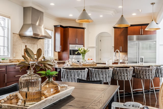 kitchen with hanging light fixtures, wall chimney exhaust hood, arched walkways, appliances with stainless steel finishes, and decorative backsplash