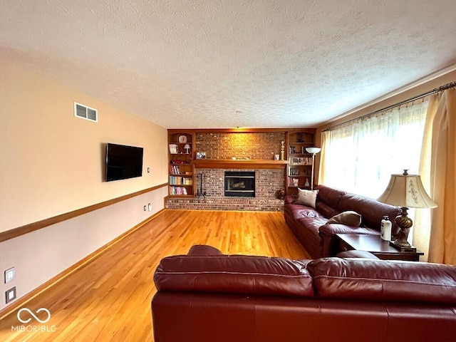 living room featuring a textured ceiling, visible vents, a brick fireplace, built in shelves, and light wood finished floors