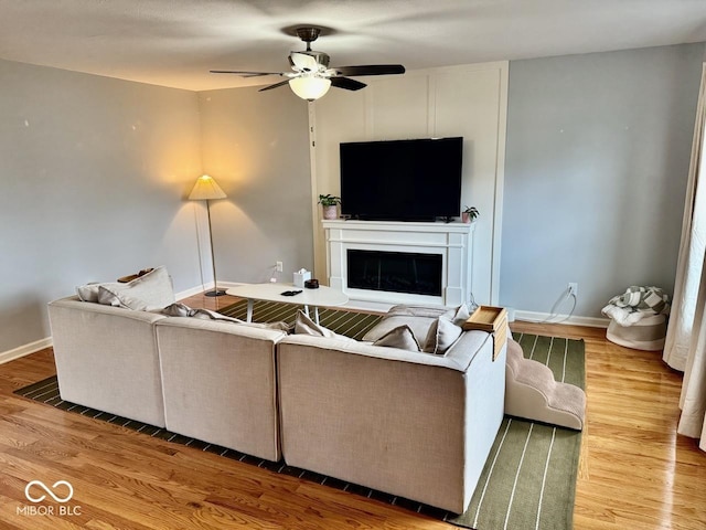 living area featuring a ceiling fan, baseboards, a glass covered fireplace, and wood finished floors