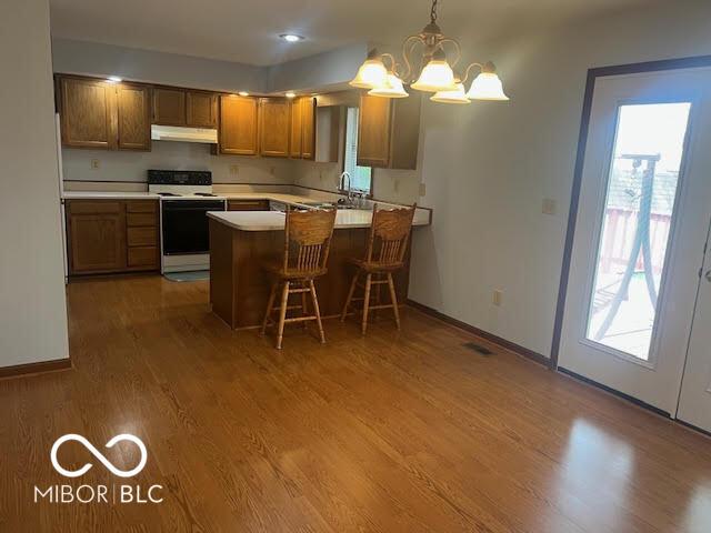 kitchen featuring a peninsula, wood finished floors, white electric range oven, under cabinet range hood, and brown cabinets