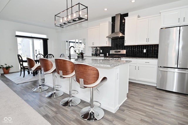 kitchen with wall chimney range hood, a kitchen island with sink, stainless steel appliances, and light wood finished floors
