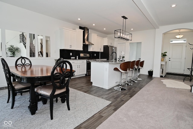 kitchen with wall chimney range hood, decorative backsplash, a kitchen island with sink, appliances with stainless steel finishes, and dark wood finished floors