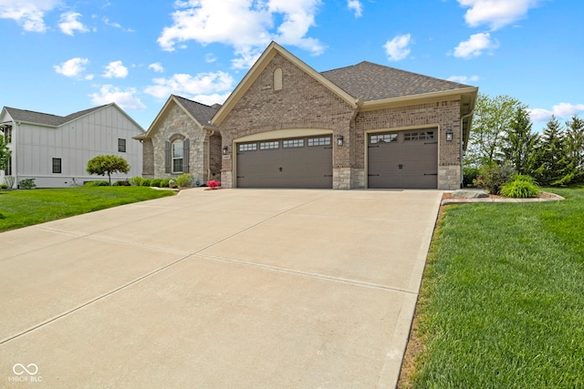 french country inspired facade featuring roof with shingles, a front lawn, stone siding, a garage, and driveway