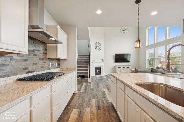 kitchen with a sink, wall chimney range hood, stainless steel gas cooktop, dark wood-type flooring, and pendant lighting