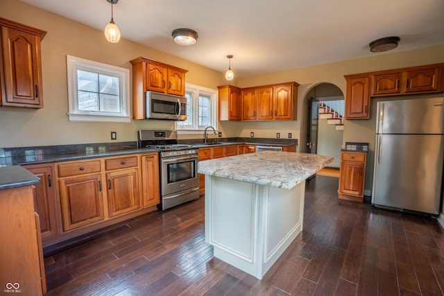 kitchen featuring appliances with stainless steel finishes, brown cabinetry, dark wood-style flooring, decorative light fixtures, and a sink
