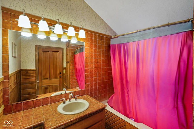 bathroom featuring wallpapered walls, shower / tub combo with curtain, tile walls, vaulted ceiling, and vanity