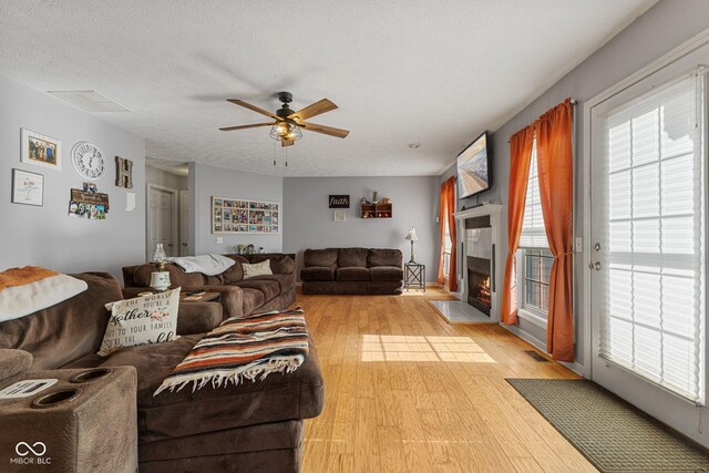 living area featuring light wood-style flooring, a ceiling fan, a fireplace with flush hearth, healthy amount of natural light, and a textured ceiling