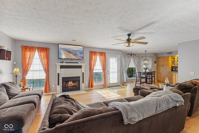 living area featuring ceiling fan, healthy amount of natural light, light wood-style flooring, a tiled fireplace, and a textured ceiling