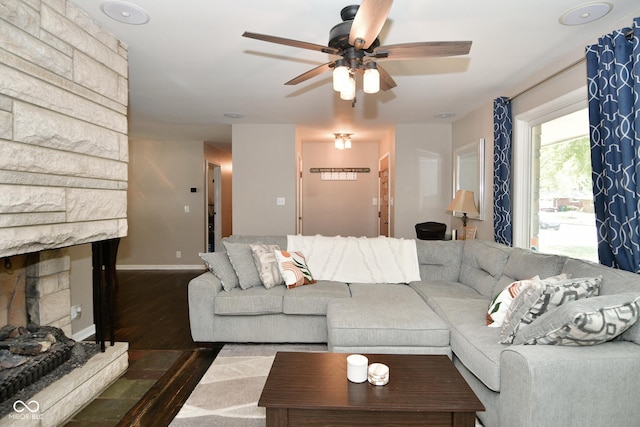 living room featuring a stone fireplace, baseboards, a ceiling fan, and dark wood-type flooring