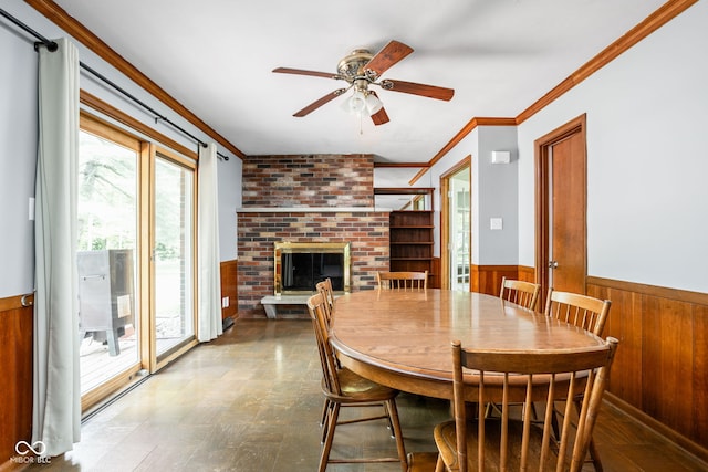 dining room with wood walls, a wainscoted wall, crown molding, ceiling fan, and a brick fireplace