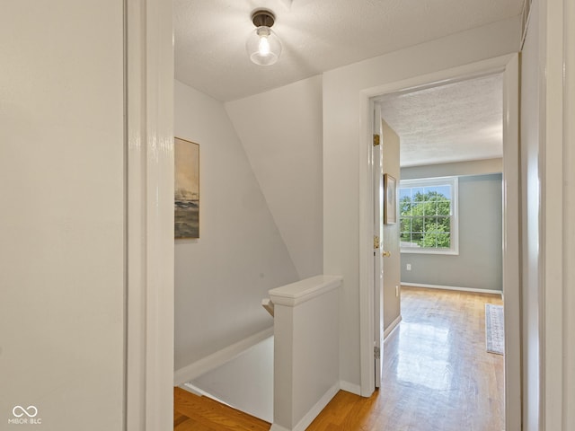 hall featuring an upstairs landing, a textured ceiling, light wood-type flooring, baseboards, and lofted ceiling